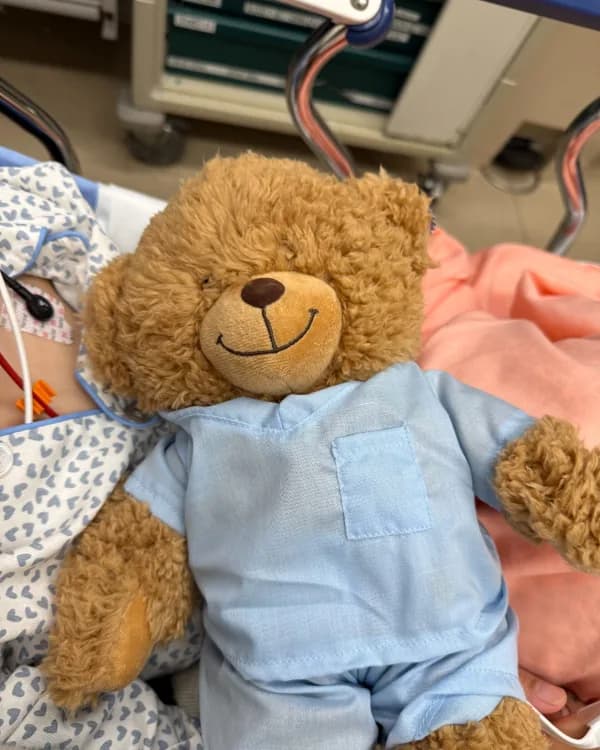 A teddy bear in blue scrubs tucked in beside D’nae’s son in a hospital bed